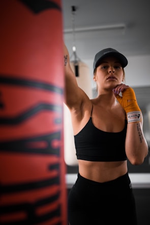 A person wearing a black sports bra and hat is in a gym setting, appearing focused and ready to punch a boxing bag. The individual has orange hand wraps and is standing in a confident pose. The background includes some gym equipment, indicating a training environment.