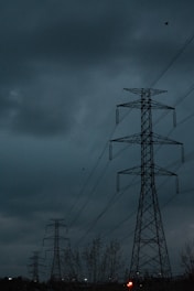 A series of tall electricity pylons in an outdoor setting beneath a dark, cloudy sky. The scene conveys an industrial feel with the strong presence of metal structures against the ominous sky.