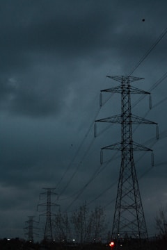 A series of tall electricity pylons in an outdoor setting beneath a dark, cloudy sky. The scene conveys an industrial feel with the strong presence of metal structures against the ominous sky.