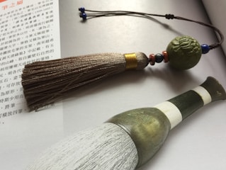 A traditional calligraphy brush with a textured, ornate handle is placed on an open book with Chinese text. The brush has a large tassel attached to a string decorated with beads and a carved spherical ornament.