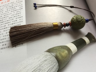 A traditional calligraphy brush with a textured, ornate handle is placed on an open book with Chinese text. The brush has a large tassel attached to a string decorated with beads and a carved spherical ornament.
