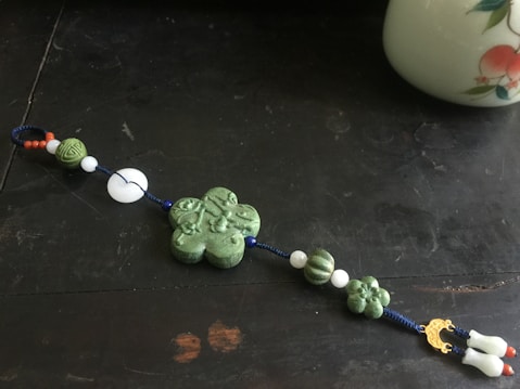 A decorative string with various beads and pendants laid on a dark surface. The central piece is a green clover-shaped bead with intricate designs. The string also includes spherical, textured beads in green, white, and a few small red ones, complemented by a gold-colored decorative element. At one end, there are two white pendants shaped like tassels. Part of a porcelain item with painted fruit is visible in the top right corner.