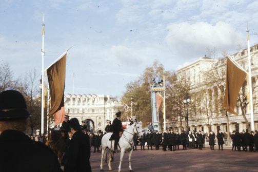 A public gathering in front of a historical building, featuring a police officer on horseback, several individuals in uniform, large flags, and spectators. The setting appears to be ceremonial, with a formal atmosphere. The sky is overcast with scattered clouds, and trees can be seen lining the area.