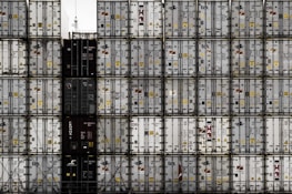 A large number of stacked shipping containers with various logos and labels, arranged in a grid pattern. The containers are primarily in shades of white, gray, and dark colors. The image gives a sense of industrial organization and mass logistics.