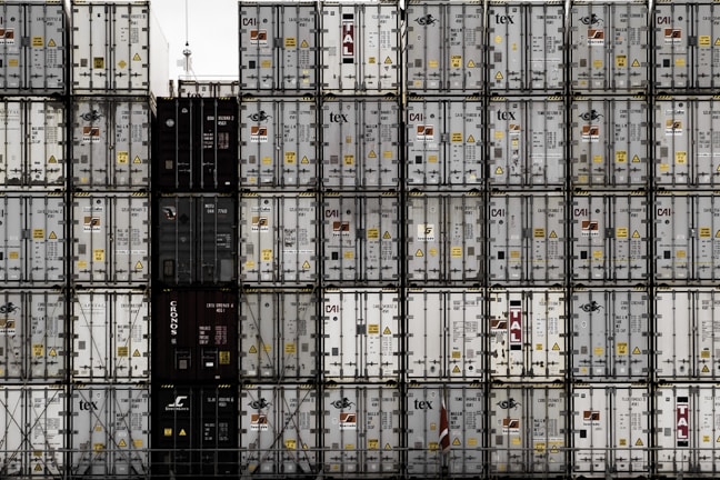A large number of stacked shipping containers with various logos and labels, arranged in a grid pattern. The containers are primarily in shades of white, gray, and dark colors. The image gives a sense of industrial organization and mass logistics.