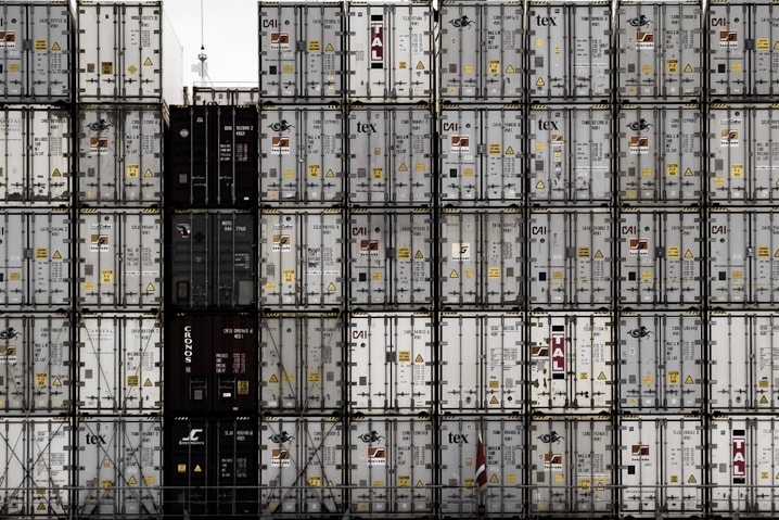 A large number of stacked shipping containers with various logos and labels, arranged in a grid pattern. The containers are primarily in shades of white, gray, and dark colors. The image gives a sense of industrial organization and mass logistics.