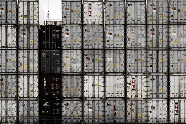 A large number of stacked shipping containers with various logos and labels, arranged in a grid pattern. The containers are primarily in shades of white, gray, and dark colors. The image gives a sense of industrial organization and mass logistics.