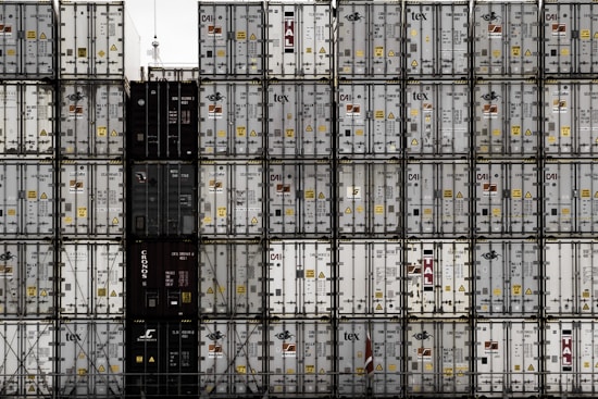 A large number of stacked shipping containers with various logos and labels, arranged in a grid pattern. The containers are primarily in shades of white, gray, and dark colors. The image gives a sense of industrial organization and mass logistics.