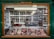 A butcher's shop window displays an array of meats neatly arranged on trays. Above the window, a sign reads 'LICENSED TO DEAL IN GAME.' Inside the shop, a couple of people are visible behind a counter surrounded by hanging overhead lights and shelves. The shop exterior features a dark green frame with wooden accents.