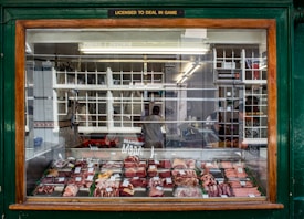 A butcher's shop window displays an array of meats neatly arranged on trays. Above the window, a sign reads 'LICENSED TO DEAL IN GAME.' Inside the shop, a couple of people are visible behind a counter surrounded by hanging overhead lights and shelves. The shop exterior features a dark green frame with wooden accents.