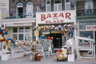 A storefront labeled 'Bazar de la Plage' is filled with a variety of colorful beach balls, toys, and beach-related merchandise. The shop's entrance is framed by numerous hanging items, including inflatable balls, buckets, and spades, creating a festive and vibrant display. The building has an old-fashioned façade with large windows and a balcony, adding a vintage charm to the scene.