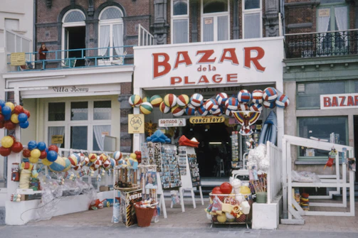 A storefront labeled 'Bazar de la Plage' is filled with a variety of colorful beach balls, toys, and beach-related merchandise. The shop's entrance is framed by numerous hanging items, including inflatable balls, buckets, and spades, creating a festive and vibrant display. The building has an old-fashioned façade with large windows and a balcony, adding a vintage charm to the scene.