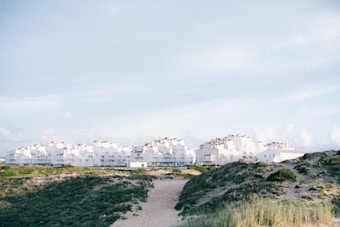 A cluster of white residential buildings is situated on a sandy, grassy hillside under a light blue sky. The architecture appears modern, with multiple stories and flat roofs adorned with small chimneys.