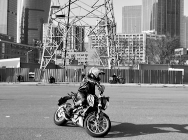 A motorcyclist wearing protective gear and a helmet rides a motorcycle on an open road. The background features a cityscape with tall modern buildings and electrical towers. Some individuals are visible in the distance, sitting and walking along the roadside.