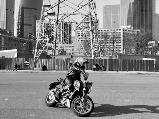 A motorcyclist wearing protective gear and a helmet rides a motorcycle on an open road. The background features a cityscape with tall modern buildings and electrical towers. Some individuals are visible in the distance, sitting and walking along the roadside.