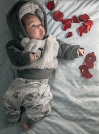 A baby dressed in a fuzzy, bear-themed hoodie and cloud-printed pants lies on a light blue, textured blanket. Red rose petals are scattered around them, adding a touch of color to the serene and cozy setting.