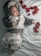 A baby dressed in a fuzzy, bear-themed hoodie and cloud-printed pants lies on a light blue, textured blanket. Red rose petals are scattered around them, adding a touch of color to the serene and cozy setting.