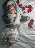 A baby dressed in a fuzzy, bear-themed hoodie and cloud-printed pants lies on a light blue, textured blanket. Red rose petals are scattered around them, adding a touch of color to the serene and cozy setting.