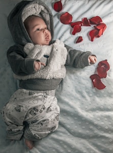 A baby dressed in a fuzzy, bear-themed hoodie and cloud-printed pants lies on a light blue, textured blanket. Red rose petals are scattered around them, adding a touch of color to the serene and cozy setting.