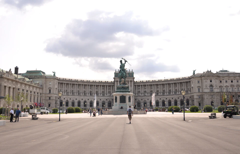 The image depicts a large, ornate building with multiple levels and columns, situated on a street corner. A statue of a man holding a sword is prominently displayed at the center of the building's courtyard. The scene is bustling with activity, featuring numerous people walking around the area and cars parked nearby.