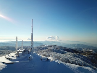 A snowy landscape featuring a radio tower and communication equipment on a mountain top, surrounded by frosted trees and distant rolling hills under a clear blue sky. The scene conveys a sense of isolation and tranquility.