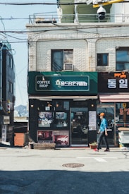 A small coffee shop with a green sign sits on a quiet street. The building is made of white bricks with wires running across the facade. A person dressed in a blue jacket and orange cap is walking past the shop. Various posters and paintings decorate the shop's exterior, and there are some potted plants near the entrance.