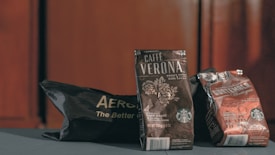 Three coffee bags are displayed in a low-lit setting. The central bag prominently features the label 'Caffè Verona' with floral designs, indicating a dark roast. Flanking it is a darker colored bag with 'Guatemala Antigua' branding and an Aeropress bag partially visible.