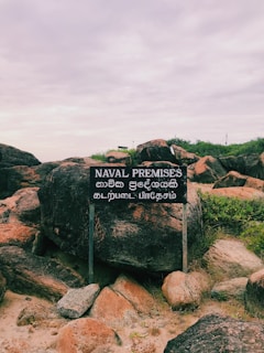 A weathered sign stands amidst a rough, rocky terrain with green shrubs growing between the large stones. The sign displays text in English reading 'NAVAL PREMISES' along with text in two other languages.