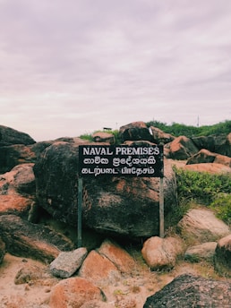 A weathered sign stands amidst a rough, rocky terrain with green shrubs growing between the large stones. The sign displays text in English reading 'NAVAL PREMISES' along with text in two other languages.
