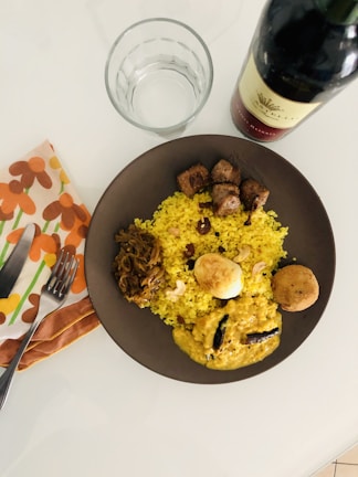 A table setting featuring a plate of vibrant yellow rice accompanied by various dishes including a curry, boiled egg, meat, and vegetables. Adjacent to the plate is a full bottle of red wine, an empty glass, a fork and knife set on a floral napkin.