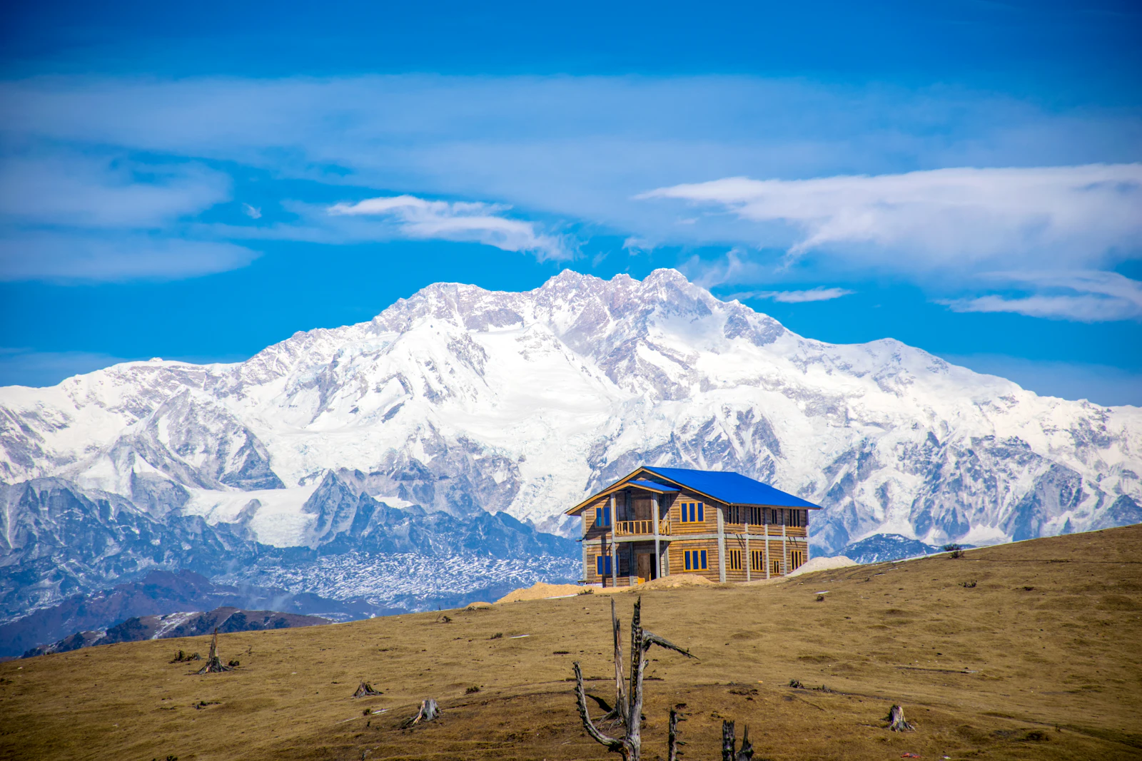 A wooden lodge on brown winter grass at Sandakphu, with the snow massif of Kanchenjunga filling the sky behind