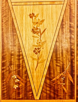 A detailed inlay design on wood featuring floral patterns. The central portion displays vertical flowers with leaves arranged in a symmetrical fashion. Surrounding the central area are two elongated floral designs, adding an artistic symmetry to the overall composition. The craftsmanship highlights intricate detailing and a smooth finish.