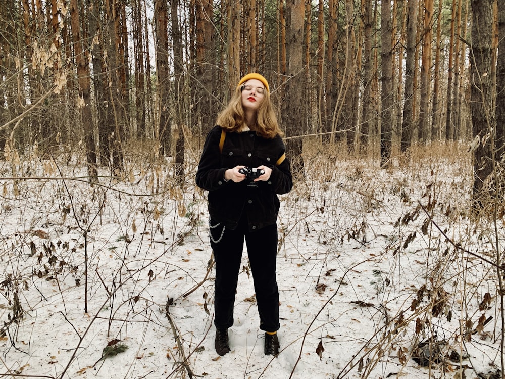 donna in giacca nera in piedi su terreno coperto di neve circondato da alberi spogli durante il giorno