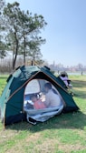 A green tent is set up on grass in a park with a clear blue sky overhead. Inside the tent, a child wearing a pink top and an adult are sitting on a blanket. Nearby, there is a baby stroller. Tall trees and a view of a city skyline in the background add to the scenic outdoor setting.