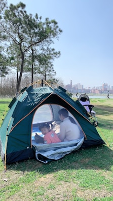 A green tent is set up on grass in a park with a clear blue sky overhead. Inside the tent, a child wearing a pink top and an adult are sitting on a blanket. Nearby, there is a baby stroller. Tall trees and a view of a city skyline in the background add to the scenic outdoor setting.