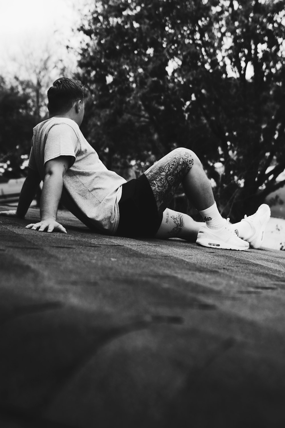 grayscale photo of boy in t-shirt and shorts sitting on concrete pavement