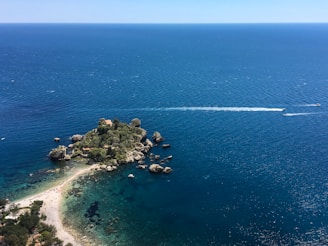 A small, rocky island covered with lush green vegetation is set against a vast expanse of deep blue ocean. Trails of white water indicate the passage of boats, creating a dynamic contrast with the serene environment. The island is connected to a sandy shore by a narrow strip of beach, surrounded by clear, turquoise waters.