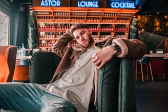 A man is sitting casually on a bar lounge chair, wearing a brown jacket and light blue jeans. The background features a bar with shelves filled with bottles and a neon sign above that reads 'Astor Lounge Cocktail'. Comfortable chairs and modern decor enhance the atmosphere.