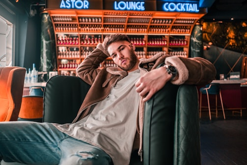 A man is sitting casually on a bar lounge chair, wearing a brown jacket and light blue jeans. The background features a bar with shelves filled with bottles and a neon sign above that reads 'Astor Lounge Cocktail'. Comfortable chairs and modern decor enhance the atmosphere.