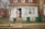 A small, urban house with a brick facade and a front door where two dogs are sitting, possibly guarding the entrance. There is a 'Beware of the Dog' sign on the door. The front yard is enclosed with a wire fence, and there are two green trash bins near the entrance. A bare tree is visible on the left side, and the surrounding area appears to be a residential neighborhood.