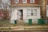 A small, urban house with a brick facade and a front door where two dogs are sitting, possibly guarding the entrance. There is a 'Beware of the Dog' sign on the door. The front yard is enclosed with a wire fence, and there are two green trash bins near the entrance. A bare tree is visible on the left side, and the surrounding area appears to be a residential neighborhood.