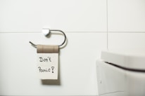 A bathroom scene displays an empty toilet paper holder with a cardboard roll attached. A piece of paper with the words 'Don't Panic!' written on it is hanging from the roll. Adjacent to the holder is a white toilet with a modern design. The background features white tiled walls, providing a clean and minimalist setting.
