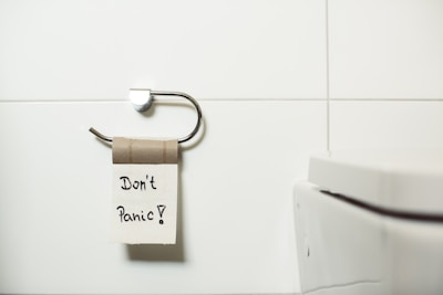 A bathroom scene displays an empty toilet paper holder with a cardboard roll attached. A piece of paper with the words 'Don't Panic!' written on it is hanging from the roll. Adjacent to the holder is a white toilet with a modern design. The background features white tiled walls, providing a clean and minimalist setting.