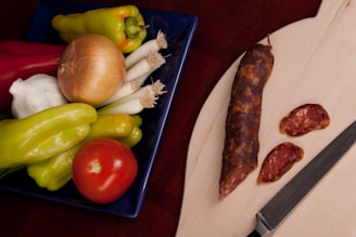 A wooden cutting board holds a whole cured sausage and two slices beside a large knife with a black handle. To the left, a blue dish contains fresh vegetables including red and yellow peppers, a tomato, a garlic bulb, an onion, and spring onions.