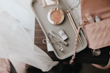 A collection of makeup items arranged on a light-colored tray on a dark surface. The tray contains a blush compact, two makeup brushes, an eyelash curler, and a few tubes of Milk makeup. A pink quilted handbag with a chain strap is partially visible next to the tray.