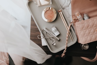 A collection of makeup items arranged on a light-colored tray on a dark surface. The tray contains a blush compact, two makeup brushes, an eyelash curler, and a few tubes of Milk makeup. A pink quilted handbag with a chain strap is partially visible next to the tray.