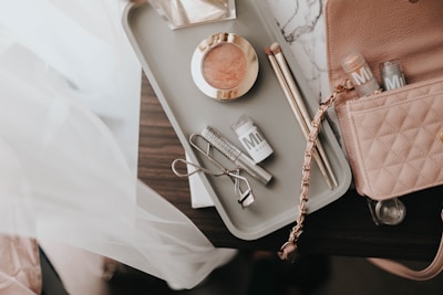 A collection of makeup items arranged on a light-colored tray on a dark surface. The tray contains a blush compact, two makeup brushes, an eyelash curler, and a few tubes of Milk makeup. A pink quilted handbag with a chain strap is partially visible next to the tray.