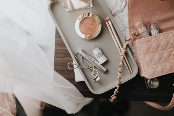 A collection of makeup items arranged on a light-colored tray on a dark surface. The tray contains a blush compact, two makeup brushes, an eyelash curler, and a few tubes of Milk makeup. A pink quilted handbag with a chain strap is partially visible next to the tray.