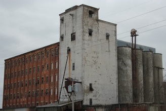 A large industrial building with weathered brick and concrete surfaces. The structure includes tall silos and an adjacent brick building with multiple windows. Power lines are visible crossing the scene.