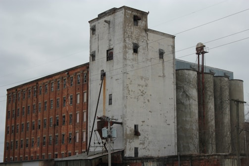A large industrial building with weathered brick and concrete surfaces. The structure includes tall silos and an adjacent brick building with multiple windows. Power lines are visible crossing the scene.